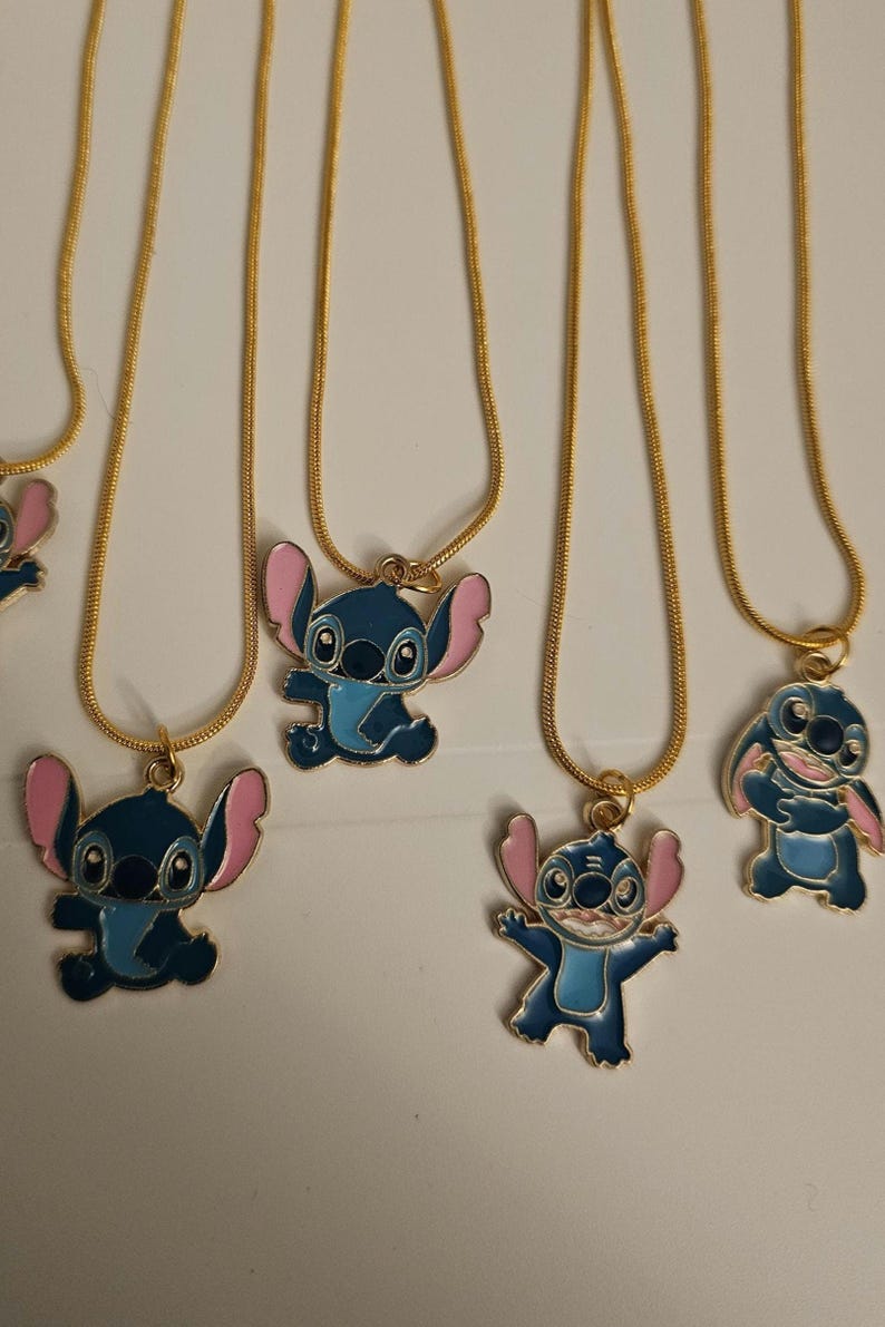 Puede incluir: Varias cadenas doradas con colgantes con el personaje de dibujos animados Stitch. Los colgantes son azules con orejas y detalles rosas. Las cadenas se muestran sobre un fondo blanco.