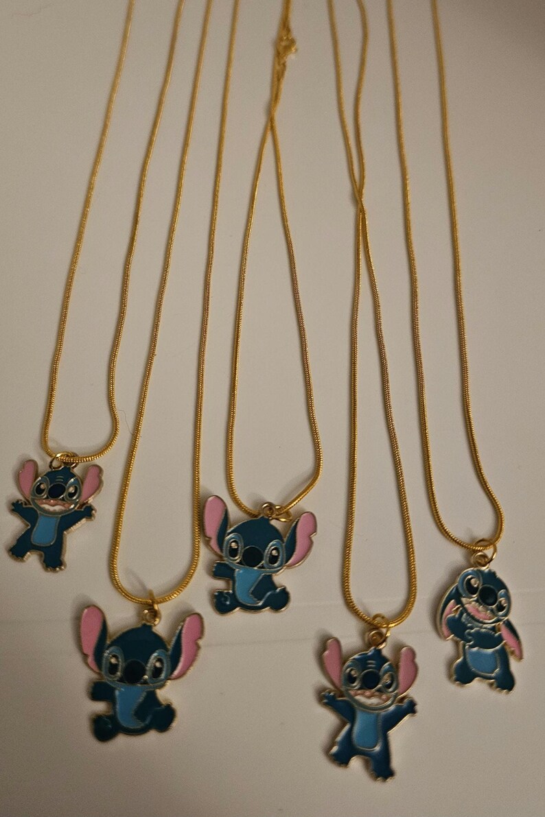 Puede incluir: Varias cadenas doradas, cada una con un colgante que representa un personaje de dibujos animados azul con orejas rosas. Los colgantes est&aacute;n en varias poses, con un cierre dorado.