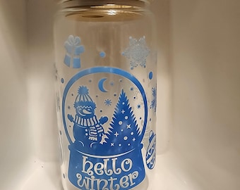 May include: A clear glass jar with a wooden lid, featuring a blue and white winter scene with a snowman, a Christmas tree, and the text "hello winter".