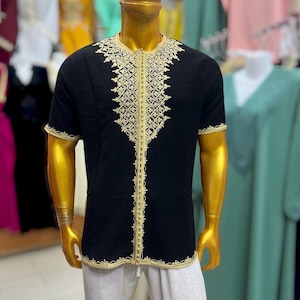 May include: A black short-sleeved tunic featuring intricate gold embroidery along the neckline, down the center front, and around the sleeve cuffs and hem. The garment has a round neck and a straight-cut silhouette, showcasing detailed metallic thread work.