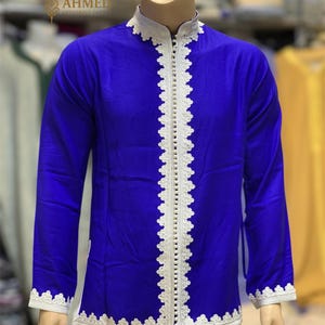 May include: A royal blue tunic with white lace trim and a mandarin collar. The tunic has long sleeves and a row of small buttons down the front. The cuffs and collar are also trimmed with white lace. The tunic is displayed on a mannequin.