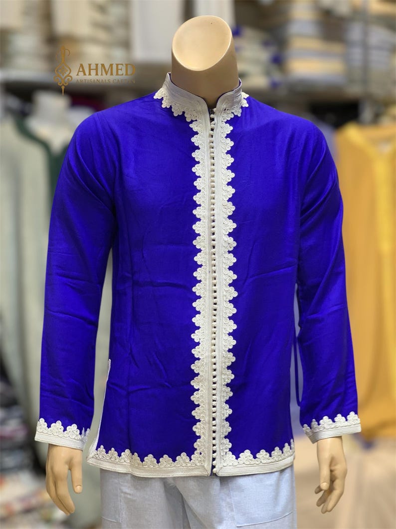 May include: A royal blue tunic with white lace trim and a mandarin collar. The tunic has long sleeves and a row of small buttons down the front. The cuffs and collar are also trimmed with white lace. The tunic is displayed on a mannequin.