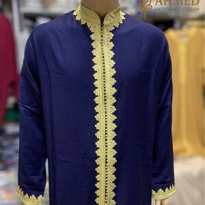 May include: A navy blue caftan with gold trim and detailing. The caftan features gold embroidery along the collar, front opening, and cuffs. The caftan is displayed on a mannequin. The text "AHMED ARTISANALS CAFTAN" is visible.