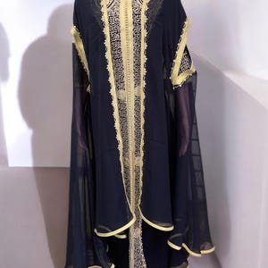 May include: A navy blue caftan with gold embroidery and trim. The caftan features a high collar and long, flowing sleeves. The garment is displayed on a mannequin. The text "AHMED ARTISANALS CAFTAN" is visible.