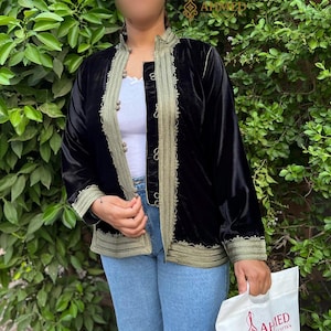 May include: A black velvet jacket featuring long sleeves, a mandarin collar, and olive green trim along the front opening, cuffs, and hem. The jacket is adorned with decorative silver-toned toggle closures and intricate embroidery along the edges.