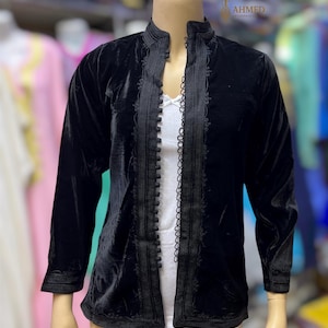 May include: A black velvet jacket featuring a mandarin collar, long sleeves, and a front opening adorned with a vertical row of small spherical buttons and intricate black braided trim detailing along the edges and cuffs, worn over a white top.