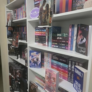 Peut inclure: Une bibliothèque blanche à plusieurs étagères remplie de nombreux livres disposés verticalement et horizontalement. Plusieurs livres sont présentés face vers l'avant sur de petits supports transparents. La collection comprend divers titres.
