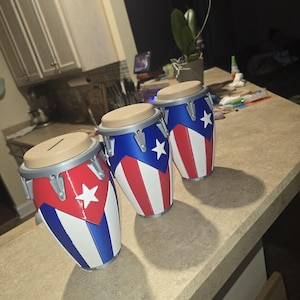 May include: Three drum-shaped coin banks featuring the design of the Puerto Rican flag. Each bank has a tan circular top with a horizontal coin slot, a blue triangular section with a white star, and vertical red and white stripes on the body.