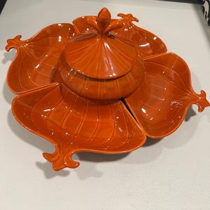 May include: An orange ceramic serving dish with a central lidded bowl and six petal-shaped compartments. The dish has a glossy finish and a decorative finial on the lid. The design is reminiscent of a flower.