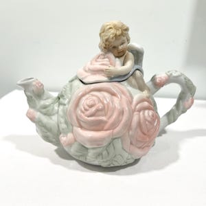 Op de afbeelding: Een porseleinen theepot met een gebeeldhouwd cherubijntje op het deksel. Het lichaam is versierd met grote, verhoogde lichtroze rozen en lichtgroene bladeren. Het handvat en de tuit zijn ook versierd met gebeeldhouwde bloem- en bladmotieven.