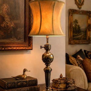 May include: A table lamp with a metallic, dark brass-toned base featuring ornate, carved detailing and a flared, warm-toned fabric shade. The lamp includes a small, side-mounted turn-key switch on the central column for operation.