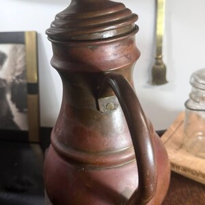 May include: A vintage copper pitcher with a rounded body, a narrow neck, and a matching lid featuring a small knob handle. The pitcher has a curved handle attached to the side and displays a weathered, oxidized patina across its metallic surface.