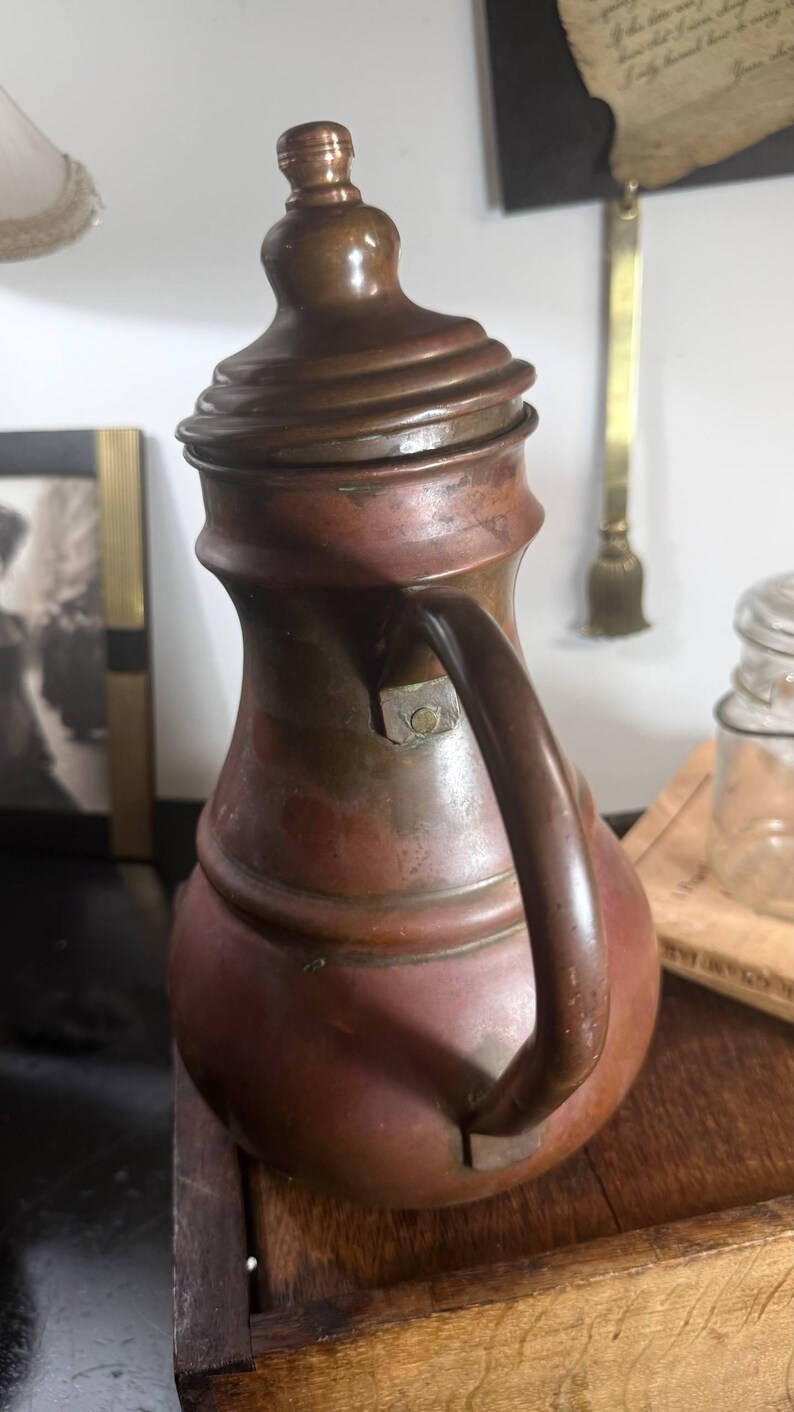May include: A vintage copper pitcher with a rounded body, a narrow neck, and a matching lid featuring a small knob handle. The pitcher has a curved handle attached to the side and displays a weathered, oxidized patina across its metallic surface.
