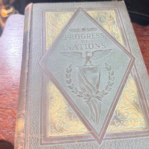May include: An antique book titled "Progress of Nations" with a green cover and gold embossed details. The cover features a diamond-shaped emblem with an eagle and text. The book is on a wooden surface.