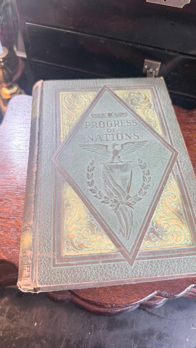 May include: An antique book titled "Progress of Nations" with a green cover and gold embossed details. The cover features a diamond-shaped emblem with an eagle and text. The book is on a wooden surface.