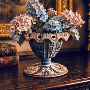 May include: A weathered, dark grey metal urn with a scalloped rim and circular cutouts, featuring a pedestal base with embossed leaf details. The urn is filled with a bouquet of blue and pink hydrangea flowers with green leaves.