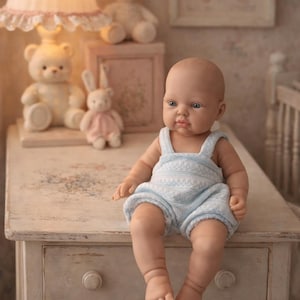 May include: A realistic baby doll with blue eyes and a smooth head, dressed in a pale blue romper. The doll is seated on a distressed white chest of drawers, with a lamp, teddy bears, and a framed picture in the background.