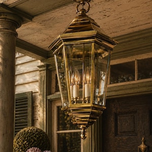 May include: A polished brass hanging lantern fixture with a hexagonal frame and clear glass panels. The light features three internal candle-style bulbs with white sleeves, suspended from a matching brass ceiling canopy by a circular ring mount.