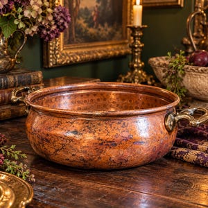 May include: A round, hammered copper pot with a wide opening and two curved brass handles attached to the sides. The exterior surface displays a weathered, mottled patina with dark brown and orange tones, while the interior shows a textured finish.