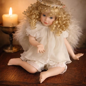 May include: A porcelain angel doll with blonde, curly hair, blue eyes, and a delicate face. The doll wears a white dress, shorts, and feathered wings. A floral halo adorns the doll's head. The doll is seated on a wooden surface.