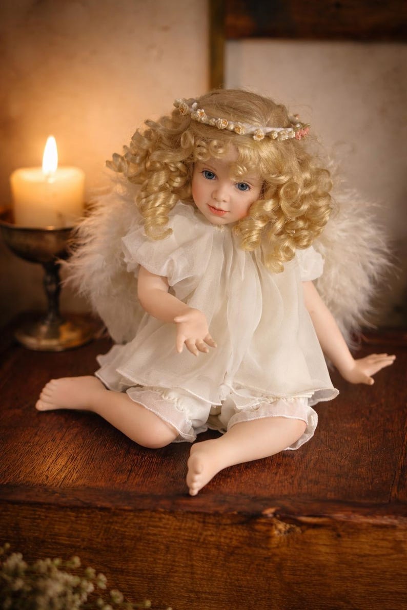 May include: A porcelain angel doll with blonde, curly hair, blue eyes, and a delicate face. The doll wears a white dress, shorts, and feathered wings. A floral halo adorns the doll's head. The doll is seated on a wooden surface.