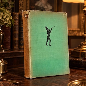 Op de afbeelding: Een hardcover boek met een vervaagde lichtgroene stoffen kaft met een zwarte silhouetillustratie van een oud-Egyptische figuur die een staf vasthoudt. Het boek heeft een versleten, beige rug met zichtbare rafels aan de bovenrand en hoeken.