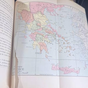 May include: A historical map of ancient Greece, displayed in an open book. The map is coloured in pastel shades, showing the different regions. The text "Ancient Greece" is visible.