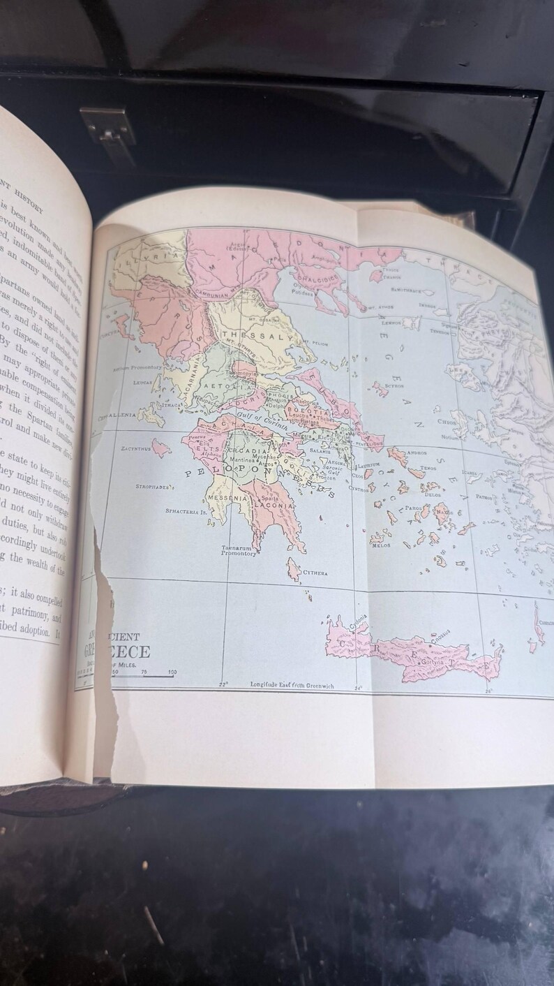 May include: A historical map of ancient Greece, displayed in an open book. The map is coloured in pastel shades, showing the different regions. The text "Ancient Greece" is visible.