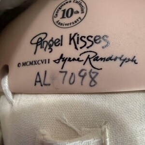 May include: Close-up of a doll's tag with the text "Angel Kisses" and "Georgetown Collection 10th Anniversary." The tag also includes the signature "Syrene Randolph" and the number "AL 7098."