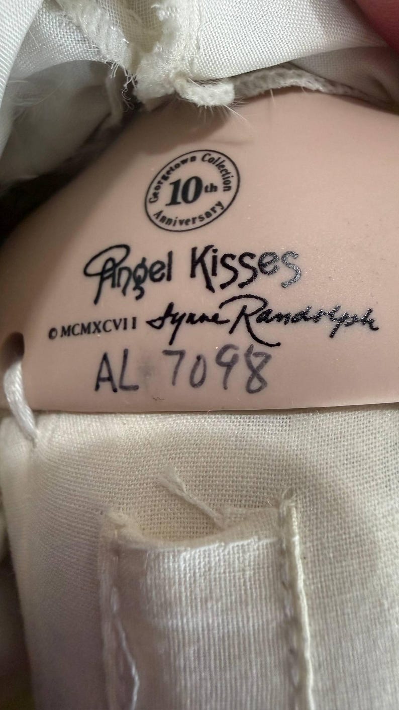 May include: Close-up of a doll's tag with the text "Angel Kisses" and "Georgetown Collection 10th Anniversary." The tag also includes the signature "Syrene Randolph" and the number "AL 7098."