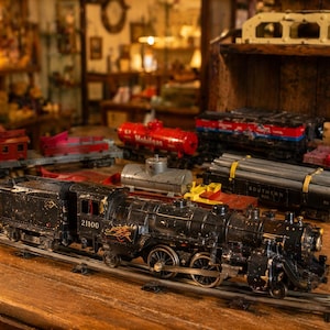 Puede incluir: Una colección de trenes de juguete antiguos, incluyendo una locomotora de vapor negra con el número 21100, varios vagones y vías. Los trenes están dispuestos sobre una superficie de madera, evocando la nostalgia y el diseño clásico de juguetes.