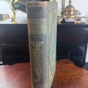 May include: An antique book titled "Progress of Nations" with a detailed embossed cover in shades of green and gold. The spine displays the title and "Volume 1". The book is set on a wooden surface, with a clock and other decorative items in the background.