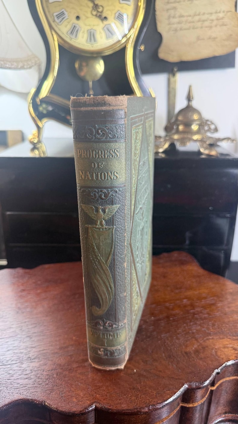 May include: An antique book titled "Progress of Nations" with a detailed embossed cover in shades of green and gold. The spine displays the title and "Volume 1". The book is set on a wooden surface, with a clock and other decorative items in the background.