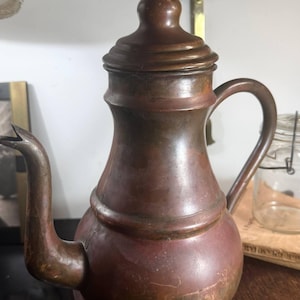 May include: A copper teapot with a rounded body, a curved spout, and a matching lid featuring a small knob handle. The metal surface displays a weathered, dark patina with visible color variations ranging from reddish-brown to deep bronze tones.