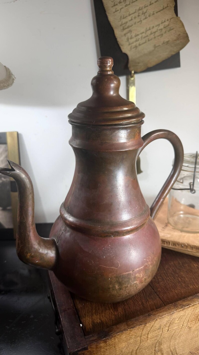 May include: A copper teapot with a rounded body, a curved spout, and a matching lid featuring a small knob handle. The metal surface displays a weathered, dark patina with visible color variations ranging from reddish-brown to deep bronze tones.
