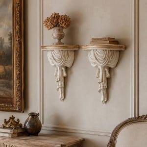 May include: Two off-white wall shelves featuring a draped fabric design with tassels. The shelves have a curved top edge with gold-toned accents. One shelf holds a decorative vase with dried flowers, and the other holds two stacked books.