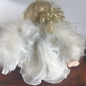 May include: A porcelain angel doll with blonde curly hair and white feathered wings. The doll is wearing a white dress with a sheer overlay. The doll is sitting on a wooden surface.