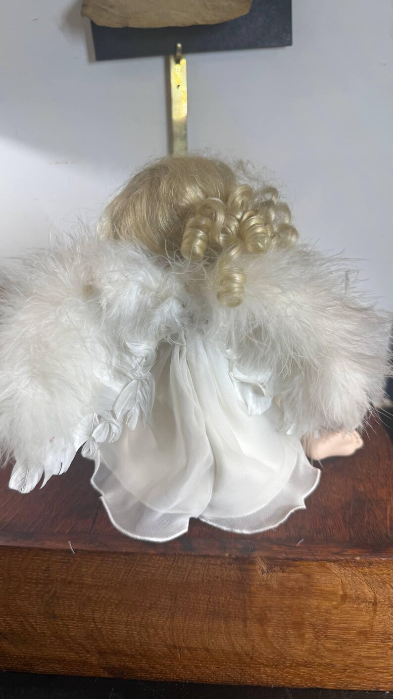 May include: A porcelain angel doll with blonde curly hair and white feathered wings. The doll is wearing a white dress with a sheer overlay. The doll is sitting on a wooden surface.