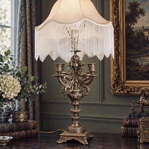 May include: A table lamp with an ornate, antique bronze-finished metal base featuring intricate scrollwork and two candle-style arms. The lamp is topped with a cream-colored fabric shade that has a scalloped edge and long, hanging white fringe.