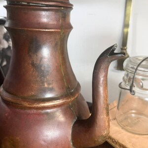 May include: A vintage copper teapot with a rounded body, a curved spout, and a matching lid featuring a small knob handle. The metal surface displays a weathered patina with visible dark oxidation marks and subtle green verdigris accents.