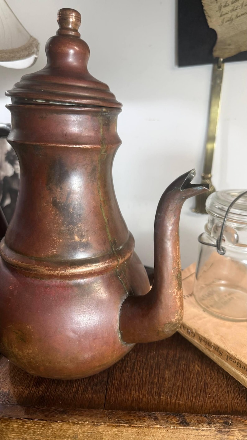 May include: A vintage copper teapot with a rounded body, a curved spout, and a matching lid featuring a small knob handle. The metal surface displays a weathered patina with visible dark oxidation marks and subtle green verdigris accents.