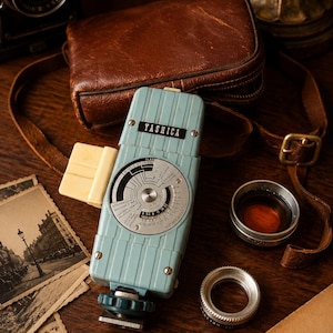 May include: A light blue Yashica light meter featuring a circular dial with black and white markings, a white side attachment, and a metal base. The device is positioned alongside a brown leather case, two circular lens filters, and vintage photographs.