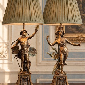 May include: A pair of table lamps featuring bronze-toned figurative bases on square pedestals. Each lamp has a pleated, sage green fabric shade topped with a decorative finial. The figures are depicted in dynamic poses with botanical accents.
