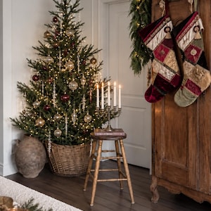 Peut inclure: Un sapin de Noël décoré dans un panier en osier, un tabouret en bois avec un candélabre à cinq bougies et deux chaussettes en velours patchwork suspendues à une armoire en bois. Les chaussettes présentent des panneaux en tissu doré, rouge et vert.