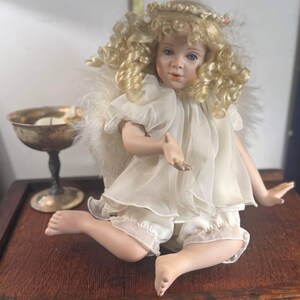 May include: A porcelain angel doll with blonde curly hair, blue eyes, and a delicate white dress. The doll is seated on a wooden surface, with a silver chalice visible in the background. The doll has feathered wings and a halo.