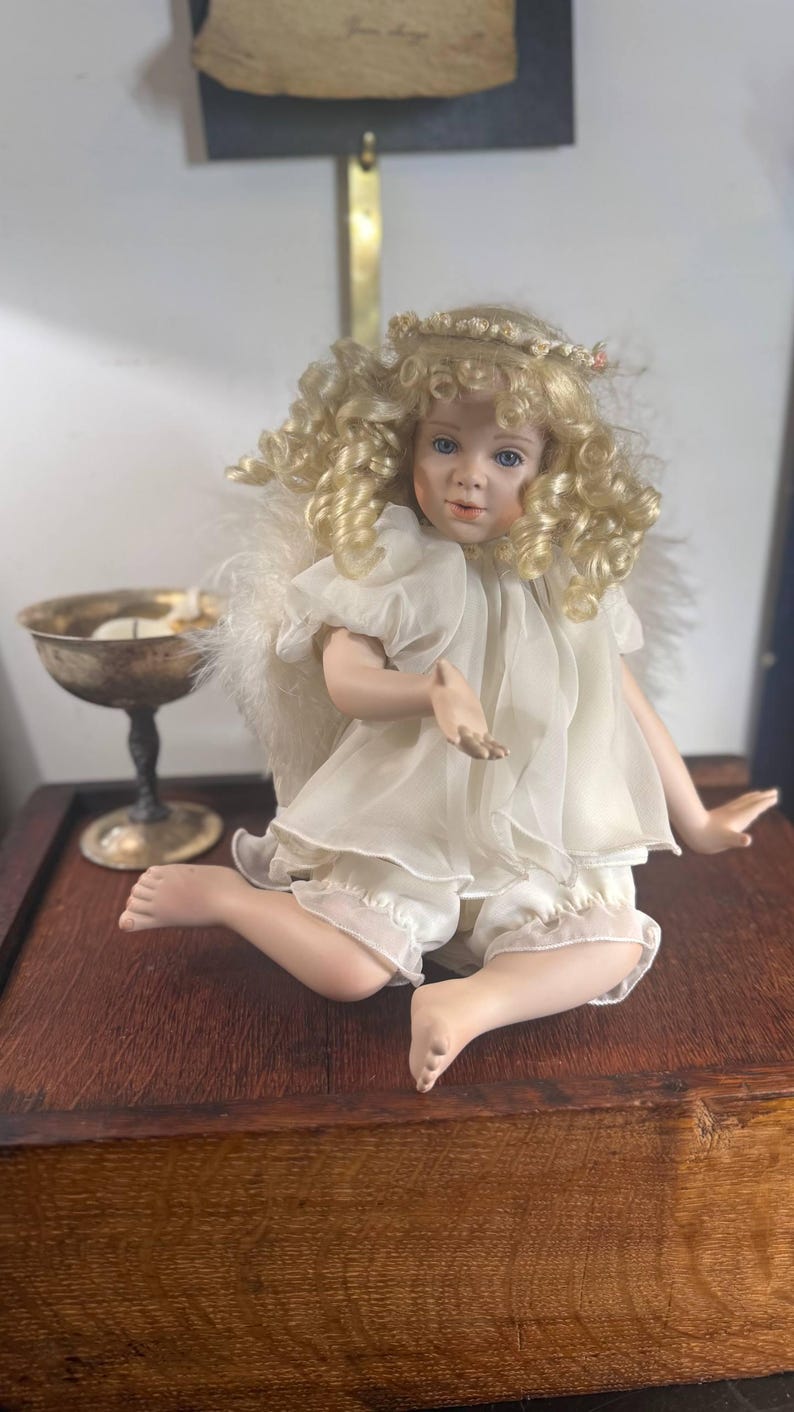 May include: A porcelain angel doll with blonde curly hair, blue eyes, and a delicate white dress. The doll is seated on a wooden surface, with a silver chalice visible in the background. The doll has feathered wings and a halo.