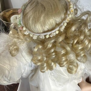 May include: A porcelain angel doll with blonde, curly hair and a delicate floral halo. The doll is wearing a white dress with feathered wings. The doll is a decorative item.