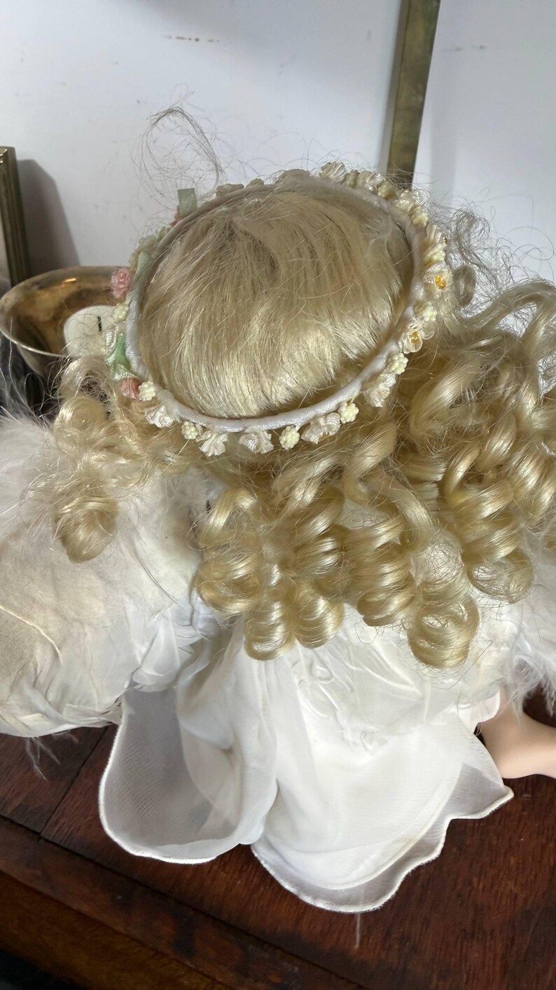 May include: A porcelain angel doll with blonde, curly hair and a delicate floral halo. The doll is wearing a white dress with feathered wings. The doll is a decorative item.