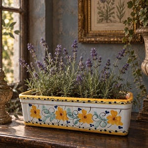 May include: A rectangular ceramic planter with a white base, featuring hand-painted yellow flowers, green leaves, and blue dots. The rim is bordered in yellow with blue accents. The planter is filled with a cluster of purple lavender stems.