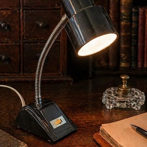 May include: A black desk lamp with a flexible, silver-colored neck and a rectangular base. The lamp is turned on, illuminating the desk. A small, clear glass dish and a leather-bound notebook are also on the desk.